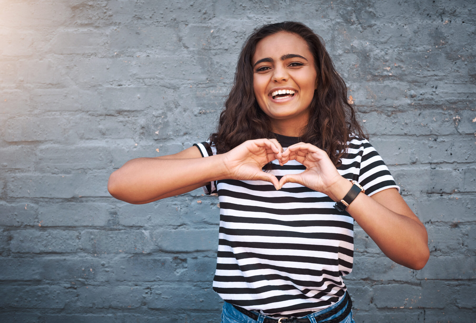 Portrait of a young woman making a heart shape with her hands against a grey wall